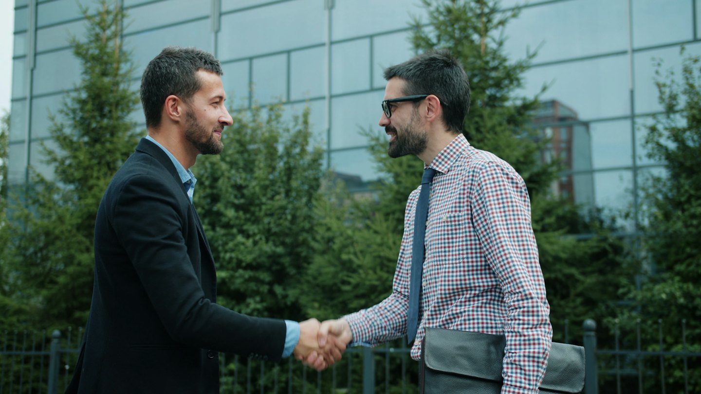 Two businessmen shake hands outdoors in front of a modern building.
