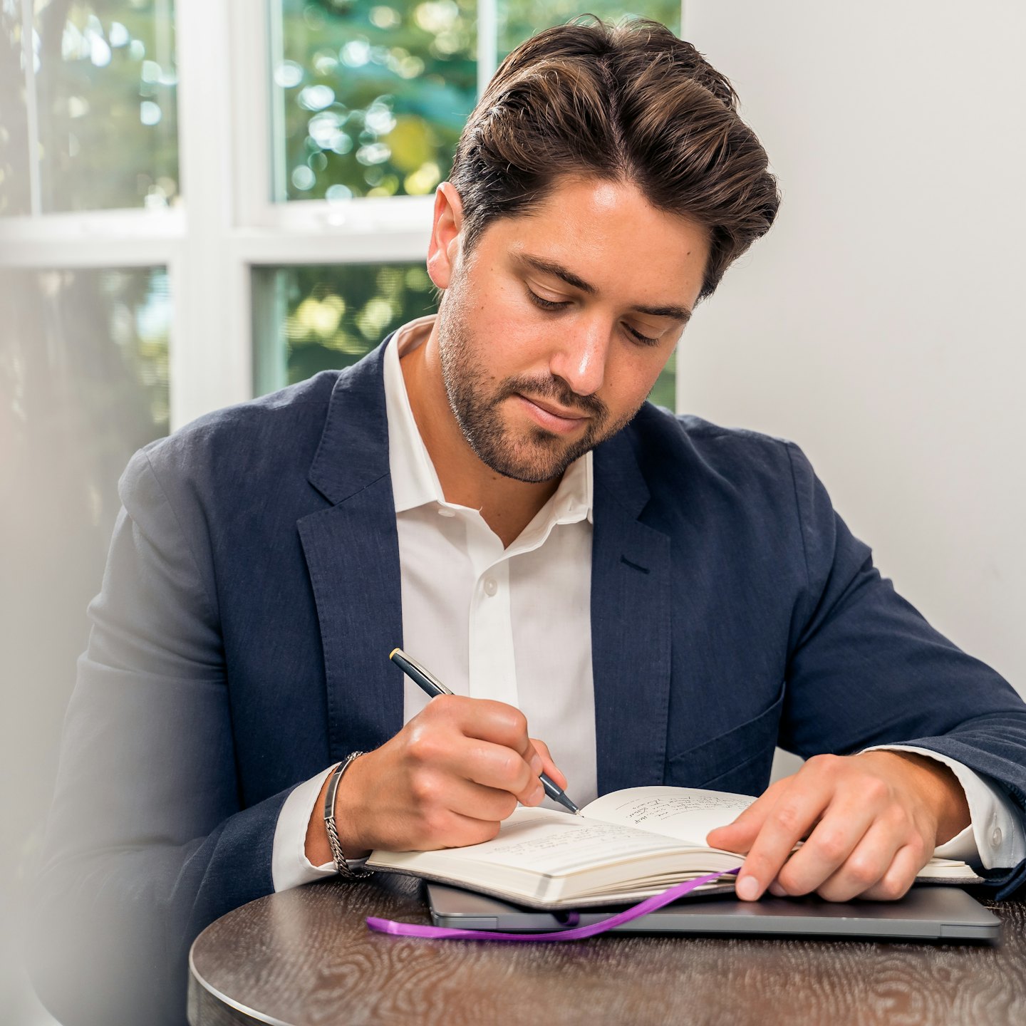 Man in blue blazer writes in notebook at wooden table.