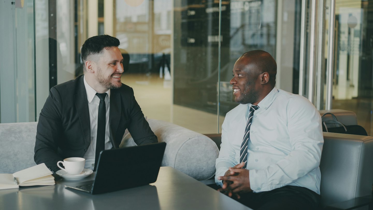 Two businessmen smiling and talking at a table in a lounge.